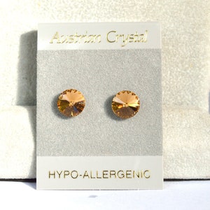 May include: A pair of gold-toned hypoallergenic earrings with amber-colored Austrian crystal studs. The earrings are displayed on a white card with the text "Austrian Crystal" and "Hypo-allergenic".