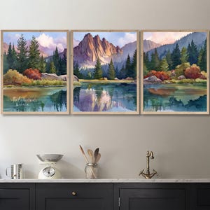 May include: A triptych of framed watercolor landscape paintings. Each features a mountain range reflected in a lake, surrounded by trees and colorful foliage. The artwork has a natural, serene theme, with a light wood frame.