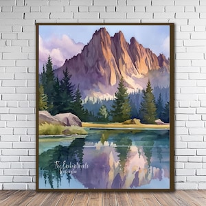 May include: Watercolor painting of a mountain landscape with a lake. The artwork features a mountain range with peaks illuminated by sunlight, reflected in the calm water. Evergreen trees and a shoreline are visible. The artwork is framed in a brown wooden frame.
