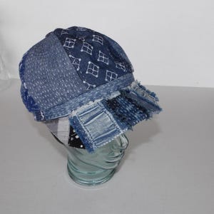 Puede incluir: Gorra de mezclilla patchwork con una visera corta. La gorra presenta varios tonos de mezclilla azul, con costuras y parches visibles. El diseño incluye un patrón de pequeñas casas blancas en el panel superior. El borde de la visera está deshilachado.