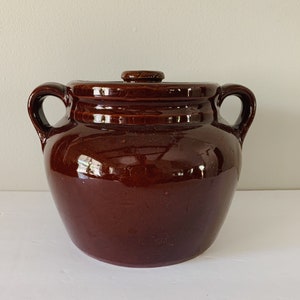 May include: A brown ceramic bean pot with a lid. The pot has two handles and a rounded body.