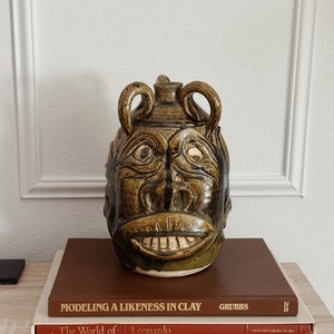 May include: A brown ceramic jug with a face sculpted on the front. The jug has two handles and a spout. The jug is sitting on top of two books. The top book has the title "Modeling a Likeness in Clay" and the bottom book has the title "The World of Leonardo".