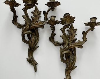 May include: Two antique bronze wall sconces. Each sconce features three candle holders and ornate, floral-inspired detailing. The sconces have a weathered, aged appearance, with intricate leaf and flower designs. The overall design is symmetrical and elegant.