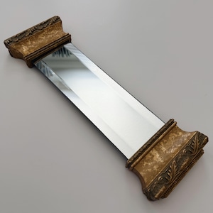 May include: A rectangular mirror featuring a beveled glass center panel flanked by two ornate, gold-toned end pieces. The decorative end caps display a textured, mottled finish with carved leaf-like detailing along the outer edges.