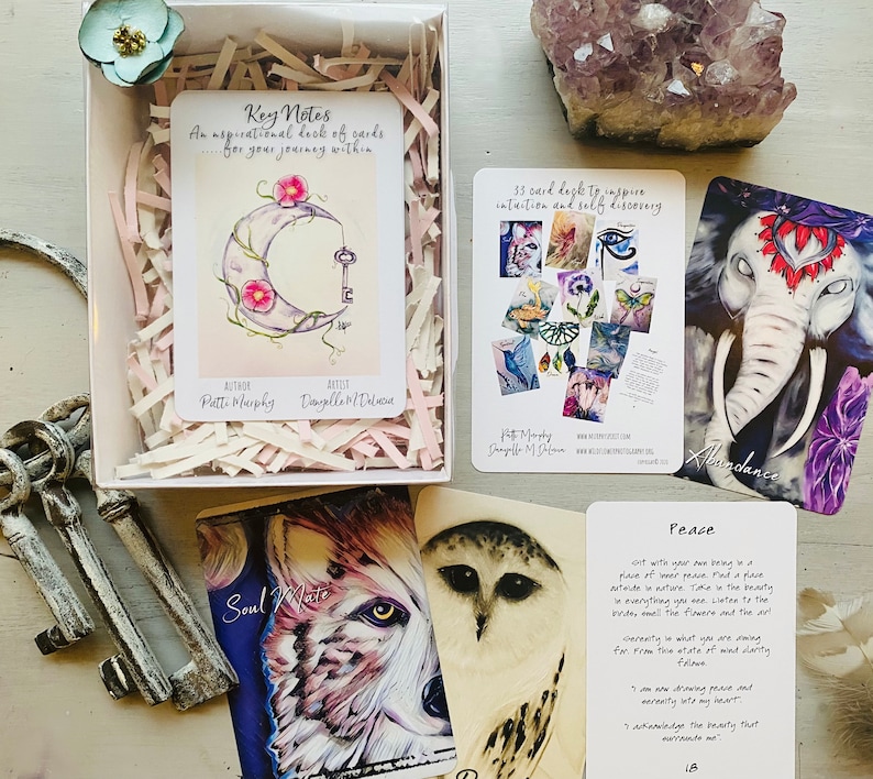 K&ouml;nnte beinhalten: Ein Set mit 35 Orakelkarten mit wei&szlig;em Hintergrund und bunten Illustrationen. Die Karten befinden sich in einer Schachtel mit rosa und wei&szlig;em Seidenpapier. Die Schachtel tr&auml;gt den Text "Key Notes: Ein inspirierendes Kartenspiel...f&uuml;r deine innere Reise." Die Karten zeigen Illustrationen eines Halbmondes, eines Schl&uuml;ssels, eines Elefanten, einer Eule und eines Wolfes. Die Karten enthalten auch Text wie "F&uuml;lle", "Seelenverwandter" und "Frieden".