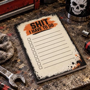 May include: A rectangular notepad with a cream-colored background, featuring a distressed orange brushstroke graphic at the top with the black text SHIT I HAVE TO DO. Below the text are ten horizontal lines with empty checkboxes on the left side.