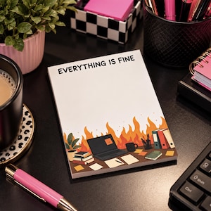 May include: A rectangular notepad with a white background and the text EVERYTHING IS FINE printed at the top. The bottom features a colorful illustration of a desk with a laptop, books, and a mug, all set against a backdrop of orange flames.