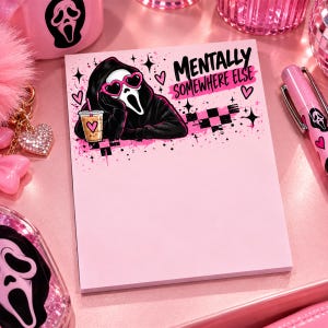 May include: A rectangular notepad with a light pink background featuring a graphic of a hooded figure wearing heart-shaped sunglasses, holding an iced coffee, and the text "MENTALLY SOMEWHERE ELSE" in black and pink lettering.