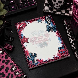 May include: A rectangular notepad featuring a white center with the text Feisty & Non Compliant. The border has a pink and blue leopard print splatter design decorated with stickers of lips, stars, a lightning bolt, a bow, a cactus, and a skull.