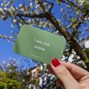 Peut inclure: Une carte verte avec le texte "Je ne suis pas seul" et les mots "The Mood Club" imprim&eacute;s en dessous. La carte est tenue dans une main sur fond d'arbre &agrave; fleurs blanches.
