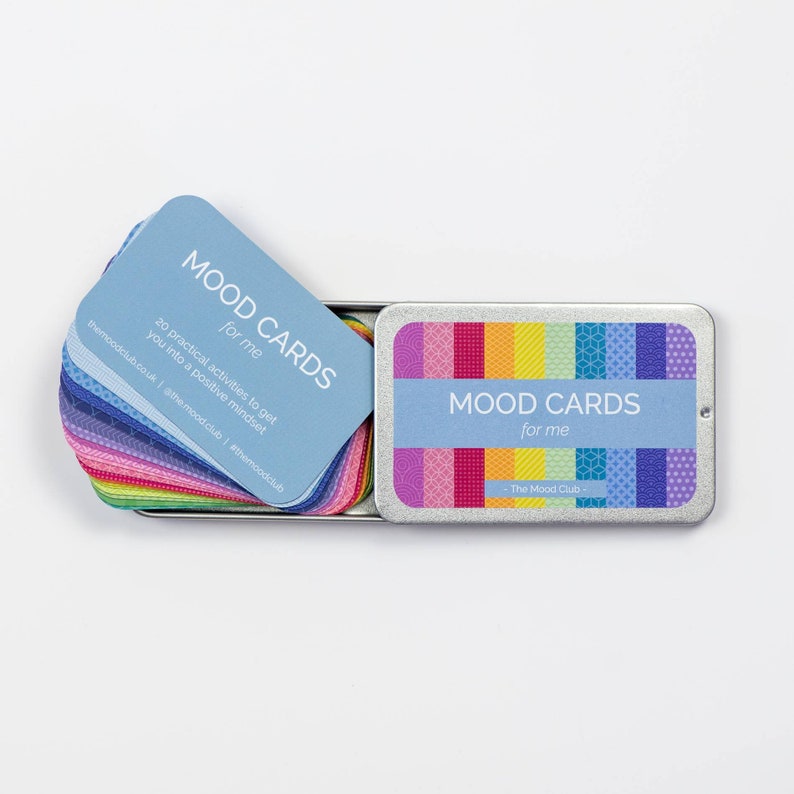 Peut inclure: Une bo&icirc;te en m&eacute;tal argent&eacute;e avec un motif arc-en-ciel et le texte "MOOD CARDS for me - The Mood Club -" sur le couvercle. La bo&icirc;te est ouverte et contient une pile de cartes bleues avec le texte "MOOD CARDS for me" et "20 activit&eacute;s pratiques pour vous mettre dans un &eacute;tat d'esprit positif" sur la carte du dessus.