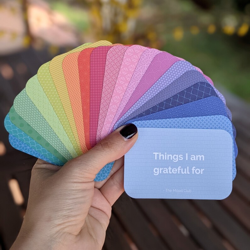 Peut inclure: Un jeu de cartes color&eacute;es avec diff&eacute;rents motifs, &eacute;tal&eacute;es dans une main. La carte du dessus est bleu clair et dit "Things I am grateful for" avec le texte "The Mood Club" en bas.