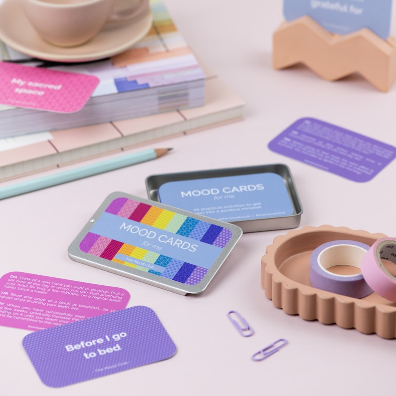 Peut inclure: Deux bo&icirc;tes en argent avec des couvercles &agrave; motifs color&eacute;s contenant des jeux de cartes d'humeur. Les cartes sont violettes et blanches avec du texte qui dit "MOOD CARDS for me" et "Before I go to bed".