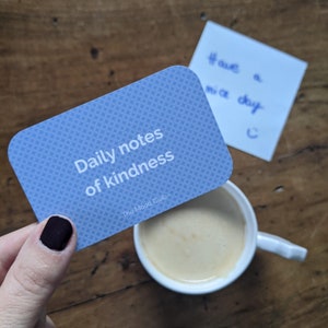 Peut inclure: Une carte bleue avec le texte "Daily notes of kindness" et le logo "The Mood Club" sur une surface en bois.