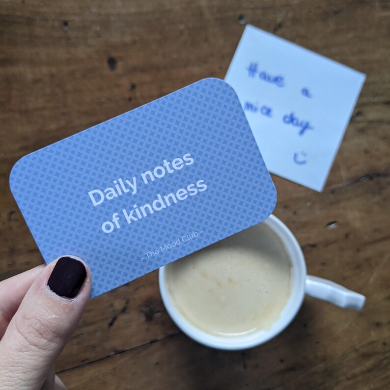 Peut inclure: Une carte bleue avec le texte "Daily notes of kindness" et le logo "The Mood Club" sur une surface en bois.