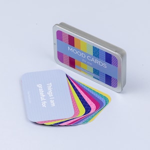 Peut inclure: Un jeu de cartes d'humeur dans une bo&icirc;te en m&eacute;tal argent&eacute;e. Les cartes sont bleues avec du texte blanc qui dit "Things I am grateful for". La bo&icirc;te a un design color&eacute; et le texte "Mood Cards for me The Mood Club".