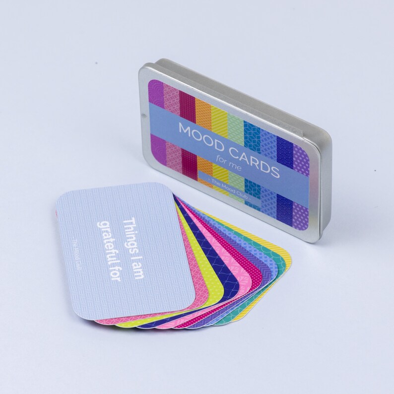 Peut inclure: Un jeu de cartes d'humeur dans une bo&icirc;te en m&eacute;tal argent&eacute;e. Les cartes sont bleues avec du texte blanc qui dit "Things I am grateful for". La bo&icirc;te a un design color&eacute; et le texte "Mood Cards for me The Mood Club".