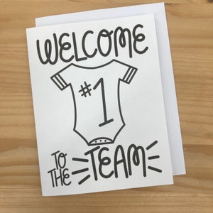 May include: A white card with black text and a black and white illustration of a baby onesie with the number 1 on it. The text reads "Welcome to the team".