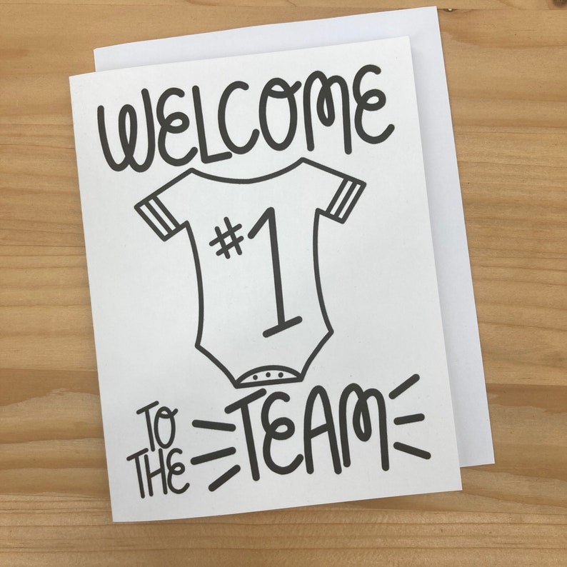 May include: A white card with black text and a black and white illustration of a baby onesie with the number 1 on it. The text reads "Welcome to the team".