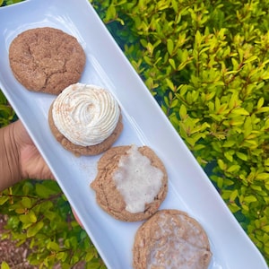 May include: A white rectangular serving tray holds four cinnamon cookies. One cookie is plain, one has a swirl of white frosting, and two are topped with a white glaze. The cookies are arranged in a row against a backdrop of green foliage.