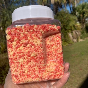Puede incluir: Un recipiente de pl&aacute;stico transparente y cuadrado lleno de una mezcla de migas rojas y beige. El recipiente tiene una tapa blanca. El contenido parece ser un producto alimenticio, posiblemente un adorno o ingrediente para postres o productos horneados.