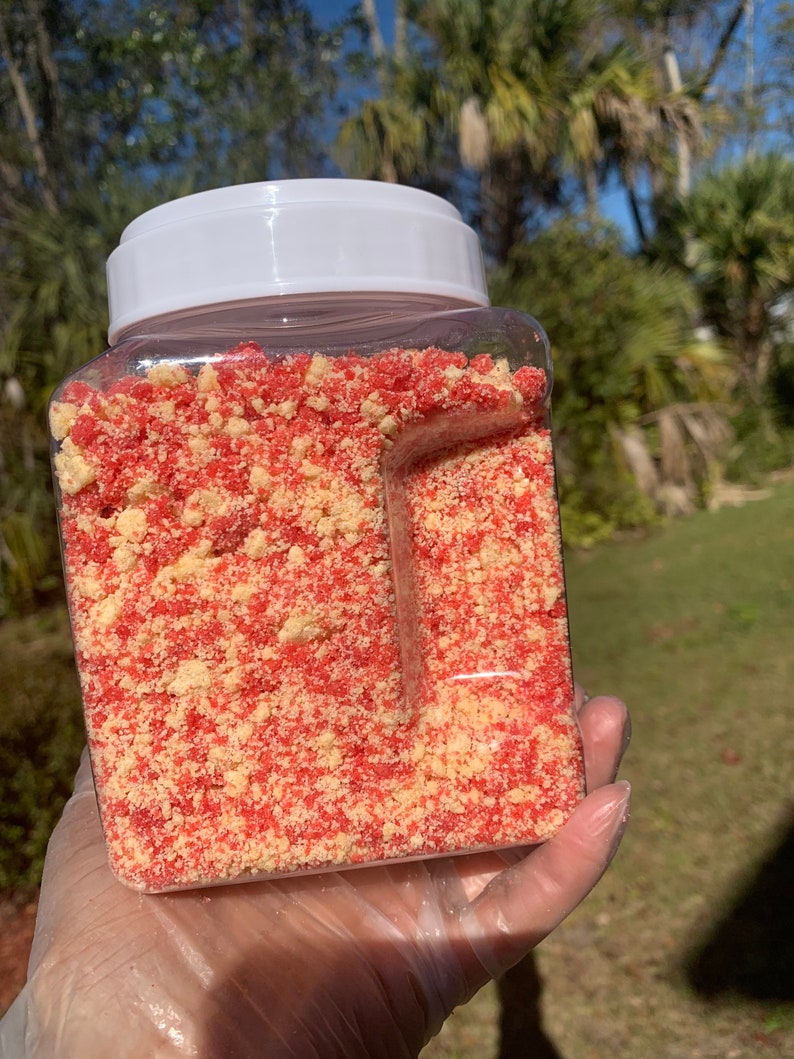 Puede incluir: Un recipiente de pl&aacute;stico transparente y cuadrado lleno de una mezcla de migas rojas y beige. El recipiente tiene una tapa blanca. El contenido parece ser un producto alimenticio, posiblemente un adorno o ingrediente para postres o productos horneados.