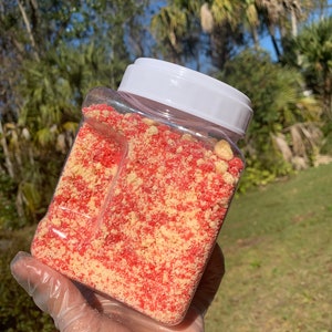Puede incluir: Un recipiente de pl&aacute;stico transparente y rectangular lleno de una mezcla de migas rojas y amarillas. El recipiente tiene una tapa blanca. El contenido parece ser un producto alimenticio, posiblemente un adorno o ingrediente para postres o productos horneados.