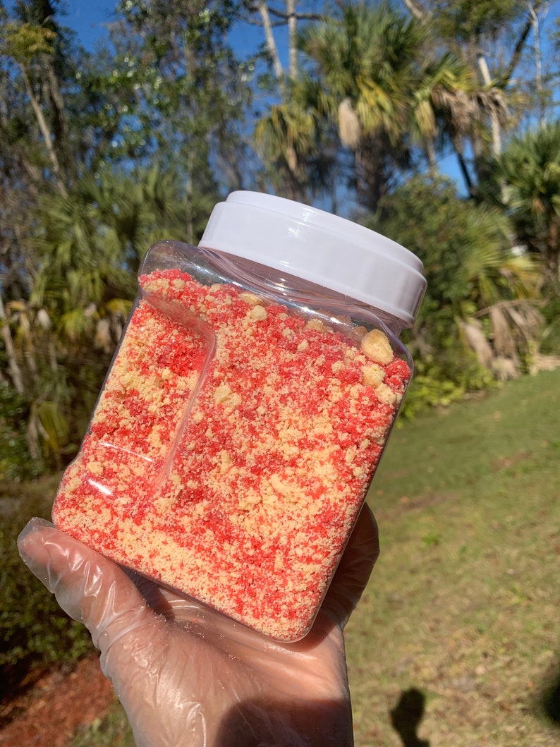 Puede incluir: Un recipiente de pl&aacute;stico transparente y rectangular lleno de una mezcla de migas rojas y amarillas. El recipiente tiene una tapa blanca. El contenido parece ser un producto alimenticio, posiblemente un adorno o ingrediente para postres o productos horneados.