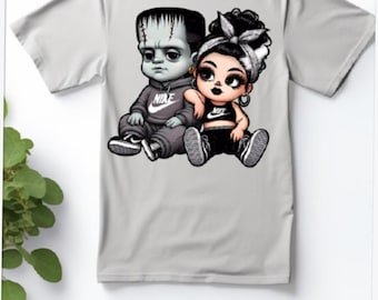 May include: A gray t-shirt with a graphic print of a cartoon couple. The male character is a Frankenstein-like monster wearing a black hoodie with a white Nike logo. The female character is a cartoon girl with black hair and a black and white Nike sports bra. The couple is holding hands and smiling.