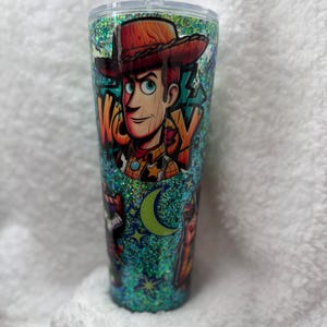 Op de afbeelding: Een glitterbeker met een cartoonafbeelding van Woody uit Toy Story. Het ontwerp bevat het woord "WOODY", een halve maan en sterren. De beker heeft een doorzichtig deksel en is gevuld met teal glitter.