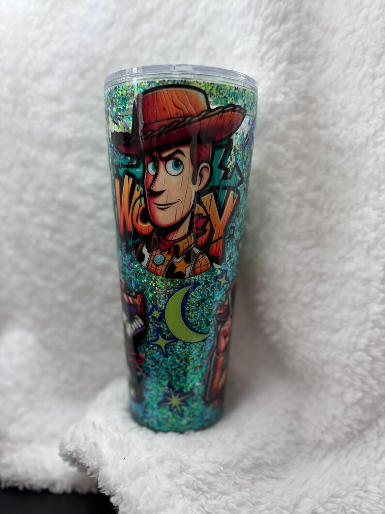 Op de afbeelding: Een glitterbeker met een cartoonafbeelding van Woody uit Toy Story. Het ontwerp bevat het woord "WOODY", een halve maan en sterren. De beker heeft een doorzichtig deksel en is gevuld met teal glitter.