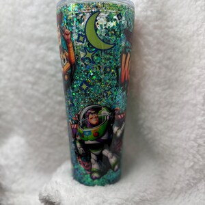 Op de afbeelding: Een hoge, doorzichtige beker met een deksel, gevuld met groene glitter en met personages uit de animatiefilm "Toy Story". Het ontwerp omvat Woody, Buzz Lightyear, een maan en sterren. De beker is cilindrisch.