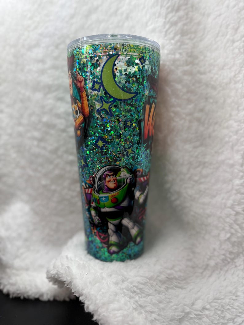 Op de afbeelding: Een hoge, doorzichtige beker met een deksel, gevuld met groene glitter en met personages uit de animatiefilm "Toy Story". Het ontwerp omvat Woody, Buzz Lightyear, een maan en sterren. De beker is cilindrisch.