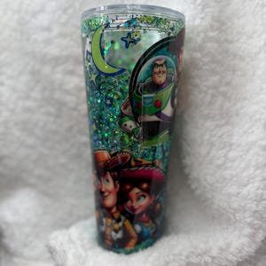 Op de afbeelding: Een doorzichtige beker met een groene glitter binnenkant en een deksel. De beker toont afbeeldingen van personages uit de film Toy Story, waaronder Buzz Lightyear, Woody en Jessie. Een maan en sterren zijn ook zichtbaar.