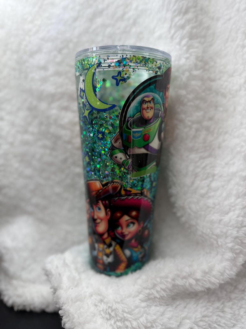 Op de afbeelding: Een doorzichtige beker met een groene glitter binnenkant en een deksel. De beker toont afbeeldingen van personages uit de film Toy Story, waaronder Buzz Lightyear, Woody en Jessie. Een maan en sterren zijn ook zichtbaar.