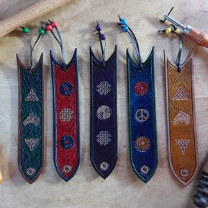 May include: A selection of handmade leather bookmarks in shades of green, red, purple, blue, and tan. Each bookmark is embossed with designs like Celtic knots, a horse, a flower, a peace symbol, and a yin-yang symbol. They are finished with beaded cords.