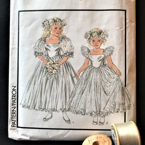 以下が含まれることがあります： ヴィンテージの縫製パターン、女の子用ドレス2着、サイズ6。パターンは、サテンのボディスとチュールスカートの白いドレス用です。ドレスはパフスリーブで、花輪で飾られています。パターンの番号は1510です。