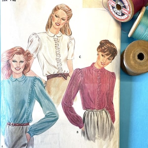 May include: A vintage Butterick sewing pattern, size 12, for blouses. The cream-coloured envelope shows illustrations of the finished garments. Spools of thread in purple, teal, and wood tones are visible in the background.