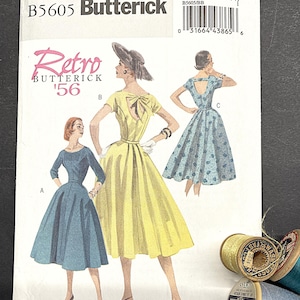 Puede incluir: Un patrón de costura Butterick vintage, B5605, con ilustraciones de vestidos retro. El patrón muestra un vestido amarillo con un lazo, un vestido azul y un vestido azul estampado. Los carretes de hilo están en primer plano.