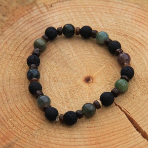 May include: A beaded bracelet featuring black lava rock beads, interspersed with green, brown, and gray stone beads. The bracelet is strung on a clear elastic cord and rests on a wooden surface.