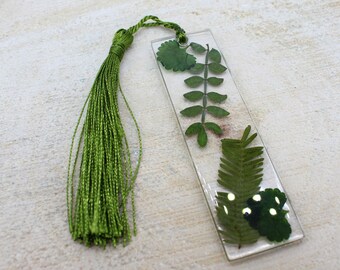 May include: A clear rectangular bookmark with pressed green leaves and a green tassel. The leaves are arranged inside the clear resin. The tassel is made of many strands of green thread.