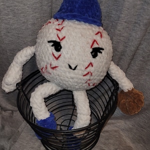 Puede incluir: Un peluche de béisbol hecho a mano con una cara sonriente, costuras rojas y un sombrero azul. El peluche tiene brazos, piernas y un guante marrón, sentado en una cesta de alambre negra. El béisbol es blanco con costuras rojas.