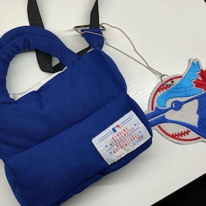 May include: A blue baseball-themed bag with a matching tag. The tag features a baseball with a blue bird and a red maple leaf. The bag has a handle and a strap. The tag is attached to the bag with a silver chain.