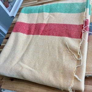 May include: A woven blanket featuring a cream-colored base with horizontal stripes in green and red. The fabric has a visible diagonal twill texture throughout the surface, and the edge of the textile shows loose, frayed white threads along the side.