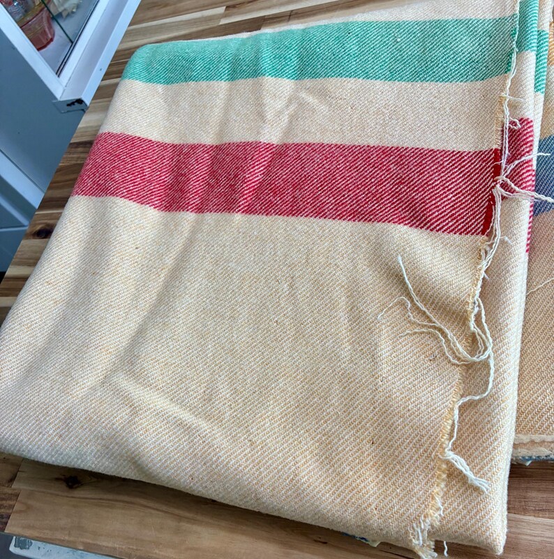 May include: A woven blanket featuring a cream-colored base with horizontal stripes in green and red. The fabric has a visible diagonal twill texture throughout the surface, and the edge of the textile shows loose, frayed white threads along the side.