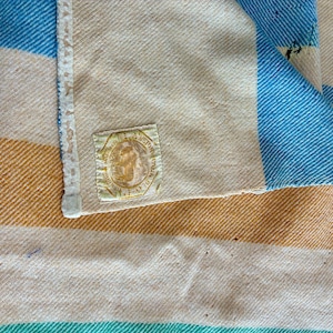 May include: A woven blanket featuring wide horizontal stripes in shades of cream, golden yellow, and bright blue. The fabric has a visible diagonal twill weave texture. A square fabric label with a circular emblem and text is stitched on.