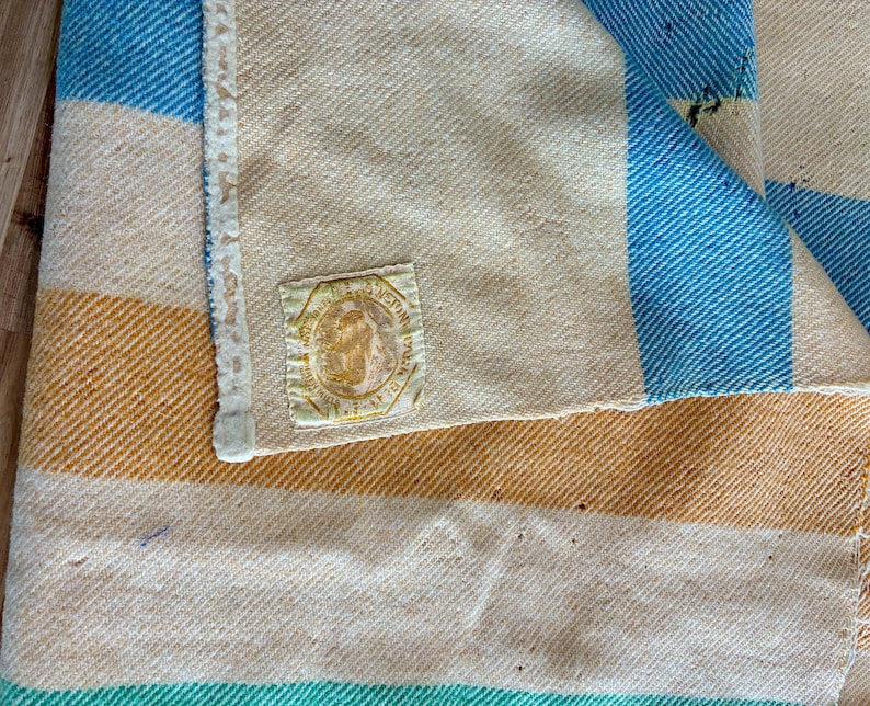 May include: A woven blanket featuring wide horizontal stripes in shades of cream, golden yellow, and bright blue. The fabric has a visible diagonal twill weave texture. A square fabric label with a circular emblem and text is stitched on.