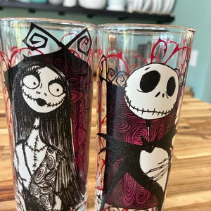 Puede incluir: Dos vasos transparentes con ilustraciones de personajes en blanco y negro. El vaso izquierdo muestra una figura femenina con cabello largo y detalles de costuras. El vaso derecho muestra una figura esquel&eacute;tica con cabeza redonda y pajarita.