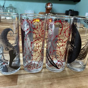 Puede incluir: Cuatro vasos de vidrio transparente con ilustraciones de personajes en blanco y negro y patrones de enredaderas rojas. Los dise&ntilde;os incluyen una figura esquel&eacute;tica, un personaje cosido y una gran forma de luna creciente en el vidrio.