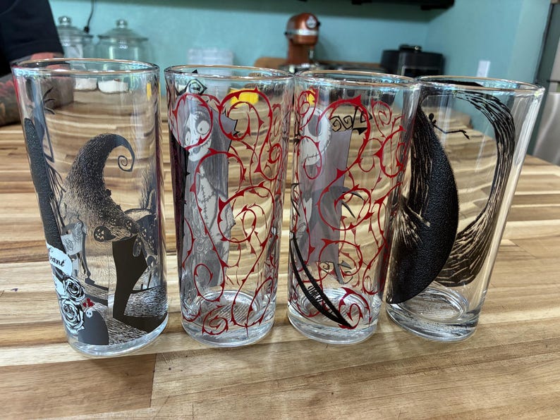 Puede incluir: Cuatro vasos de vidrio transparente con ilustraciones de personajes en blanco y negro y patrones de enredaderas rojas. Los dise&ntilde;os incluyen una figura esquel&eacute;tica, un personaje cosido y una gran forma de luna creciente en el vidrio.